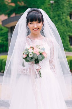 Gelin çok güzel, beyaz bir elbise giymiş ve elinde bir buket çiçek tutan bir peçe var..