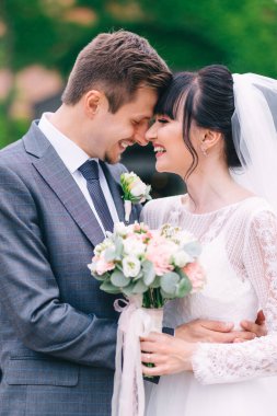 Uzun peçeli beyaz elbiseli tatlı kıvırcık saçlı gelin, şehirdeki sokakta siyah kravatlı gri takım elbiseli damadı kucaklıyor. Yeni evlilerin düğün portresi.