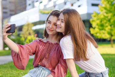 İki liseli kız, yazın bir parkta, akıllı telefondan selfie çekiyorlar, günlük kıyafetler giyiyorlar, okuldan ve üniversiteden bir süre uzaklaşıyorlar.,
