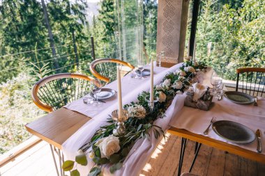 Güzel masa, sandalyeler, bardaklar, tabaklar, mumlar ve doğanın arka planına karşı bir buket çiçek. Restoran, kutlama..