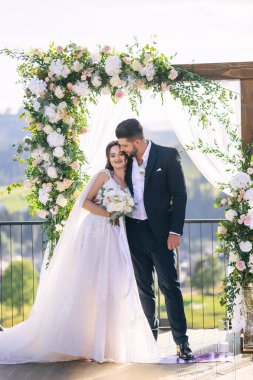 Kemer ve doğanın arka planında yeni evlilerin düğün portresi. Törende, uzun dantelli beyaz elbiseli güzel kıvırcık saçlı bir gelin ve siyah takım elbiseli şık bir damat sarılıyor..