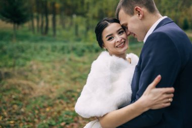 Neşeli gelin ve damat, sonbahar parkında yürürken sarılıp gülümserler..