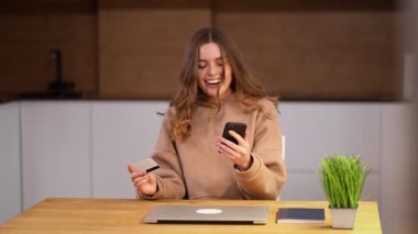 Pozitif bir genç kadın akıllı telefonu ve banka kartıyla online alışverişleri öder ve şık tasarımcı mutfağında oturur. Kız mutluluktan başını sallıyor ve ikramiye kazanıyor..
