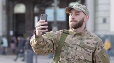 Askeri üniformalı genç bir adam istasyonda duruyor, video iletişimiyle konuşuyor, gülümsüyor..