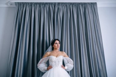 Duvağı olan güzel bir kız ellerini belinden tutuyor, gri perdelerin arkasına bakıyor. Dantelli romantik bir elbise giyiyor..