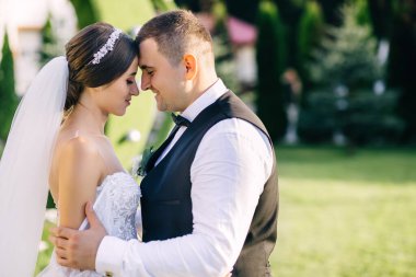 Gelin ve damat sıcak bir yaz gününde güzel yeşil bir bahçede duruyorlar. Yeni evlilerin güzel öpücüğü