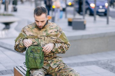 Kamuflaj üniformalı genç bir asker bankta oturur, çantasını kontrol eder, uzun bir hizmetten sonra evine döner.
