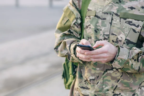 hands of a man holding a mobile phone. A soldier in a camouflage suit and a bag is typing text messages on a smartphone on a city street