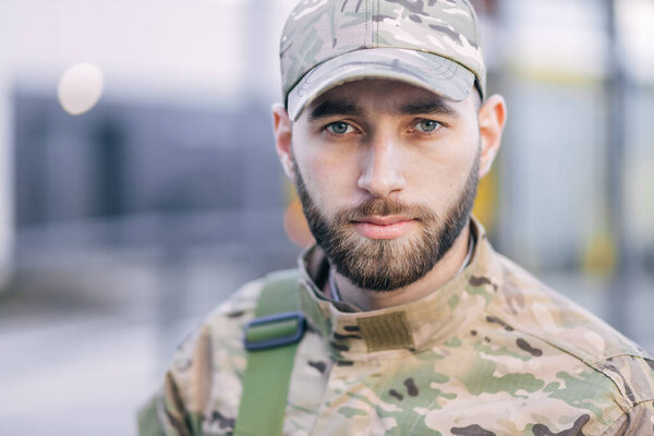 a serious young soldier standing in the street, wearing a cap and special clothing in green color.