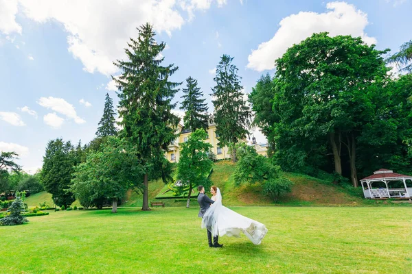 身穿黑色西服的时髦新郎和身穿白色长裙 头戴面纱的漂亮新娘在公园的绿草上跳舞 — 图库照片