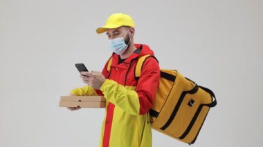 Tıbbi maskeli bir teslimatçı elinde pizza kutuları olan bir sırt çantası olan beyaz bir telefona bakıyor.