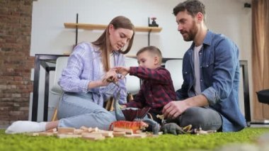 Mutlu genç anne ve baba, yeşil zeminde oturan oğluyla oyun oynuyor. Dinazorlar ellerinde. Evde oyuncaklarla ve tahta bloklarla eğleniyorlar.