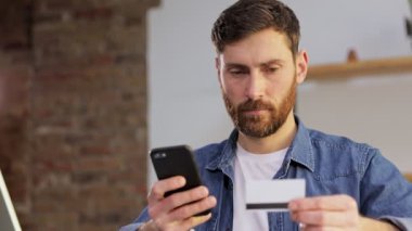 Genç bir adam evde bir masada oturuyor ve çalışıyor. Kredi kartı bilgilerini akıllı telefonuna kaydediyor. Masada bir dizüstü bilgisayar var..