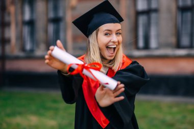 Üniversitenin arka planında tek başına duran bir öğrenci, mutlu bir şekilde diplomasını gösterir ve eğitiminin sonunu kutlar..