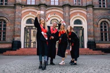Hafıza için fotoğraf. Bir grup mutlu öğrenci mezuniyet belgesiyle selfie çekiyor. üniversite binasının arka planına karşı bornozlarla.