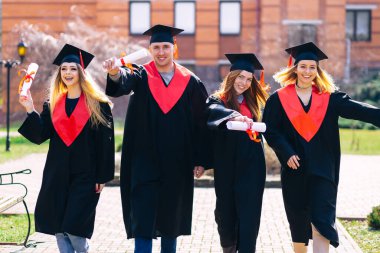 Bir grup gülümseyen üniversite mezunu ellerinde diplomalarla yürüyüp dışarıda mutlu mesut mezuniyetlerini kutluyorlar..