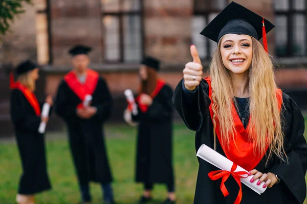 Education theme of happy graduate student outdoors on friends background. She holds a diploma, and shows a thumb up, recommends a university for students.
