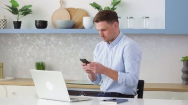 Yürekli genç adam evdeki masada bir sandalyede otururken akıllı telefon kullanıyor. Adam internette geziniyor, video izliyor, sosyal ağlar kullanıyor..