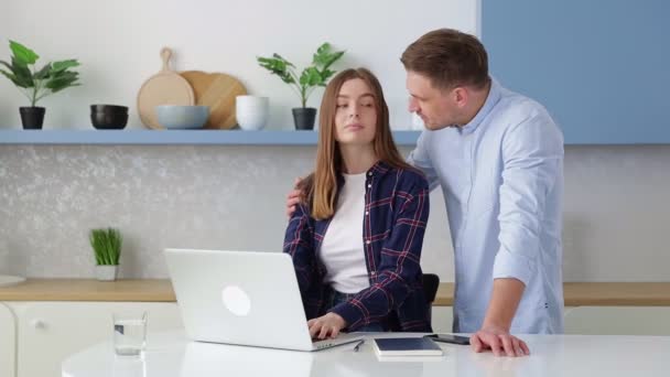 一个年轻的女人坐在一个现代化厨房的桌子旁 在笔记本电脑前工作 她的丈夫走近她 拥抱她 他们一起讨论这个项目 女孩在屏幕上展示了一些东西 — 图库视频影像