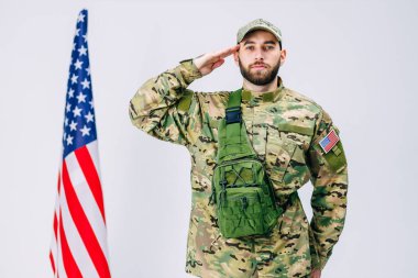 Askeri üniformalı yakışıklı askerin beyaz arka planda stüdyoda Amerikan bayrağının yanındaki kameraya bakması.