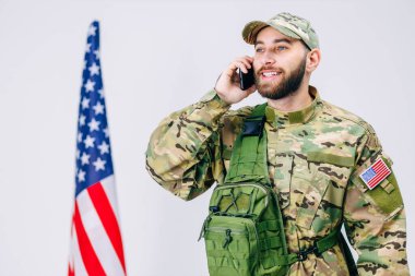 Kamuflaj üniformalı bir asker, sırt çantası, telefonda mutlu bir şekilde konuşuyor, iyi haberler alıyor, Amerikan bayrağının yanında duruyor..