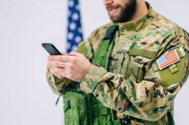 Şapkalı ve sırt çantalı mutlu asker beyaz arka planda Amerikan bayrağı yakınında yapılan askeri eğitimden sonra akıllı telefon ekranına gülümsüyor.