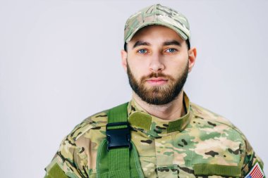 Askeri üniformalı ciddi bir askerin beyaz bir arka planda stüdyoda Amerikan bayrağının yakınındaki kameraya bakışını yakından izleyin.