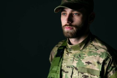 Kamuflaj kıyafetli düşünceli genç asker karanlık arka plana bakıyor.