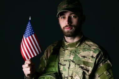 Askeri üniformalı ciddi bir asker, Amerikan bayrağını elinde tutuyor. Kameraya bakıyor. Stüdyoda karanlık arka planda kendinden emin ve ciddi.