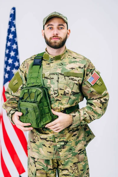a man of the USA soldier, with a serious face in a military uniform with a backpack, near the American flag, on a gray background.