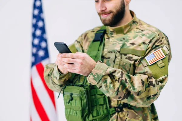 Şapkalı ve sırt çantalı mutlu asker beyaz arka planda Amerikan bayrağı yakınında yapılan askeri eğitimden sonra akıllı telefon ekranına gülümsüyor.