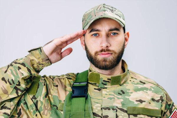 Portrait of a US Army soldier in camouflage uniform, saluting, looking at the camera. The soldier holds his hand to his head as a sign of respect.