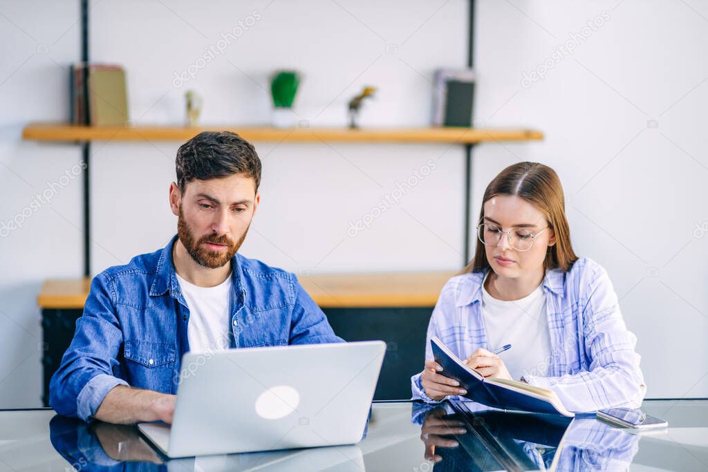 A freelance married couple working from home. A guy and a girl in casual clothes with gadgets are working in the kitchen at the table.