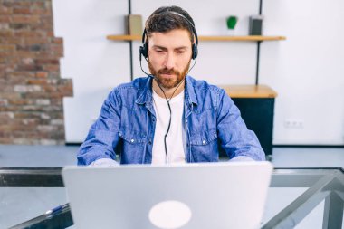 Ofiste dizüstü bilgisayarla çalışan kulaklıklı, olgun sakallı adam..