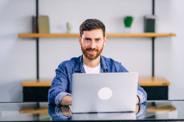 Modern mutfakta dizüstü bilgisayarda çalışan gülümseyen genç adam, e-postaları kontrol eden, sosyal ağda mesaj yazan, internet bankacılığı hizmetini kullanan mutlu genç adam, bilgi arayan