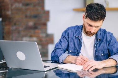 Dizüstü bilgisayarın başında oturan sakallı iş adamı defterine notlar alır. Evden çalış..