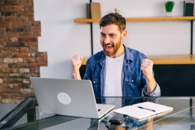 Açık tişörtlü neşeli, sakallı bir adam masada oturuyor. Kariyer konsepti geliştiriyorum. O, avantajlı bir sözleşme imzalayarak bir başarı jesti yapar. Dizüstü bilgisayarı olan bir proje üzerinde çalışıyoruz..