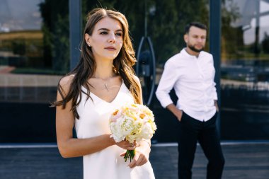 Ön planda güzel düşünceli bir gelin, elinde bir buket çiçekle, arkasında bir adam, arka planda, cam kapılar