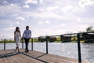 Birbirine aşık genç bir çift, bir erkek ve bir kadın, su kenarındaki ahşap iskelede yürüyorlar, el ele tutuşuyorlar ve mutlu hissediyorlar. Siyah gözlük tak..