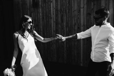 Bir buket çiçekli güzel gelin ve damat ahşap bir duvarın yanında el ele tutuşuyorlar. Aşıkların düğün portresi ve gülümseyen yeni evliler.