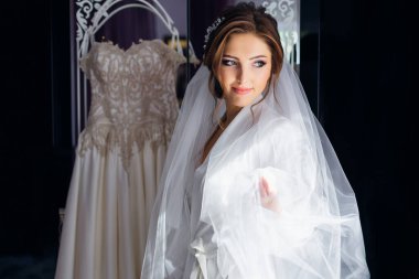 Duvak takmış sabahlık giymiş bir gelin askıda gelinliğin yanında duruyor. Günaydın bornozlu genç bayan..