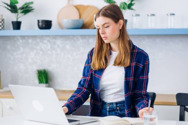 Gündelik giysiler içinde genç ve çekici bir kadın mutfakta oturmuş dizüstü bilgisayar klavyesine konsantre bir şekilde daktilo yazıyor. Güzel bir iş kadını. Uzaktan kumandalı bağımsız çalışıyor.