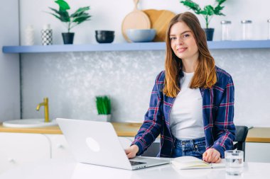 Evdeki bilgisayarda çalışan mutlu, serbest çalışan genç bir kadın. Çekici bir iş kadını çevrimiçi çalışıyor. Kameraya bakmak için dizüstü bilgisayar kullanıyor.