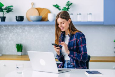 ... bilgisayarlı dizüstü bilgisayar ve akıllı telefon tarayıcıları kullanarak mutfak arka planında oturan çekici genç bir kadın. Yazılı mesaj, ekrana bakıp gülümsüyor.