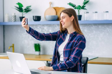 Mutlu ve çekici bir kadın, evde oturmuş akıllı telefon kullanıyor. Ailesiyle konuşuyor, iyi haberler veriyor, pozitif düşünüyor, selfie çekiyor..