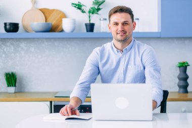 Genç serbest çalışan bir adam mutfakta arka planda oturuyor, evden çalışıyor, serbest çalışıyor. Erkek iş modeli dostça gülümsüyor..