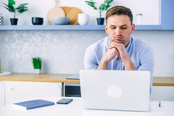 Pensive man sitting at table in modern kitchen looking at laptop pondering problem solution, pensive guy working from home