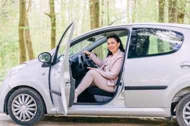 Mutlu kadın gri arabasında oturuyor yeşil bir parkta, kapısı açık..