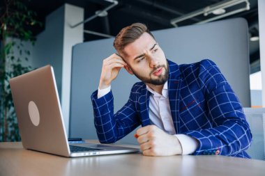 Pensive male freelancer using laptop and wireless internet in the office. The concentrated young man looks to the side.