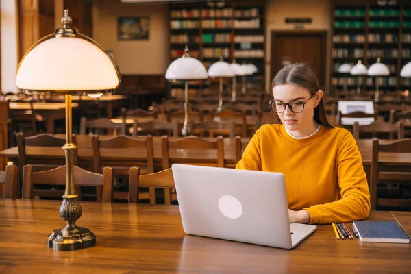 Teenage girl college student using laptop computer writing on computer while working from library or studying on technical device in class sitting at table. Online education, e-learning concept.